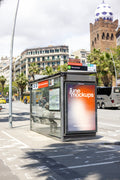 Photorealistic Barcelona bus shelter poster mockup photographed at the Monumental public transport stop on Carrer de la Marina in the Fort Pienc neighborhood of Eixample, Spain. A vertical JCDecaux city light advertising display features a colorful orange, purple and white JuneMockups gradient design, integrated into a modern glass bus shelter with a dark metal frame, visible route information and realistic reflections. Behind the advertising panel stands La Monumental, Barcelona's historic former bullring,