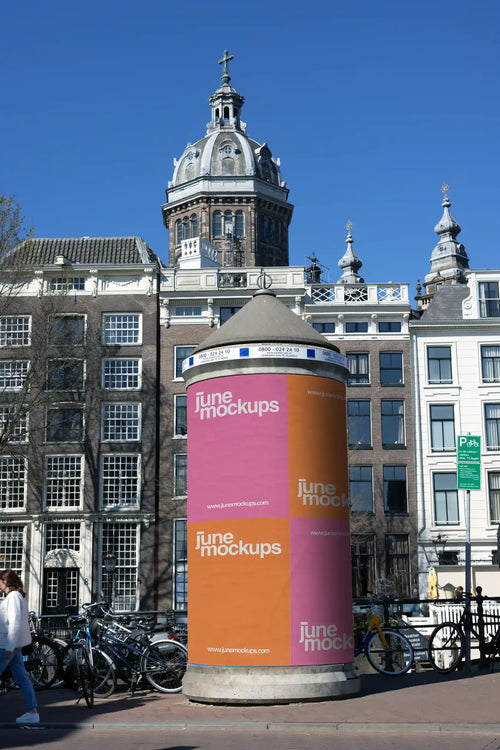 Cylindrical billboard mockup in central Amsterdam with historic architecture and St. Nicholas Basilica in the background.