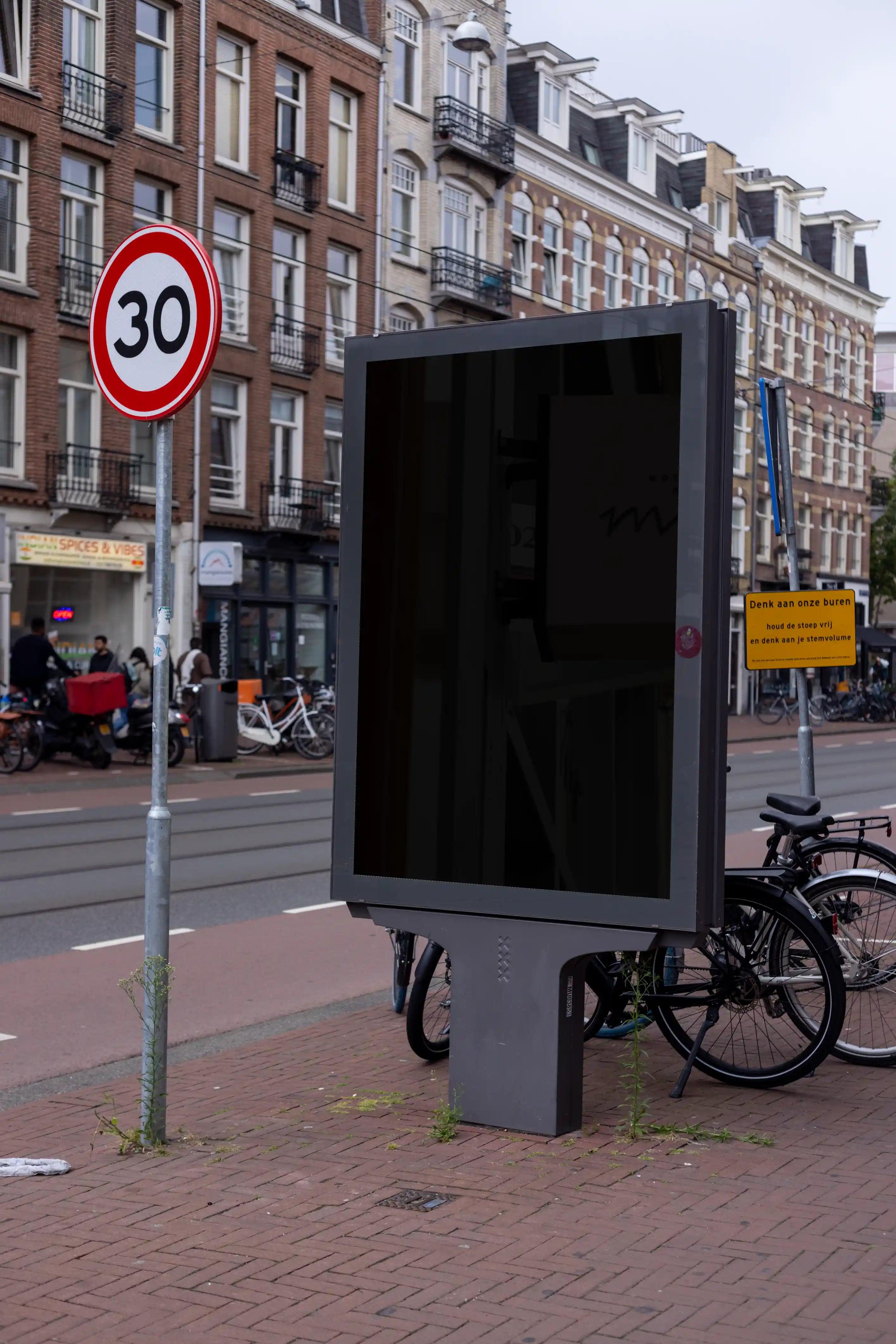 Blank screen Amsterdam De Pijp city light poster mockup placed on a busy urban street with bicycles and traditional Dutch buildings in Amsterdam, Netherlands.