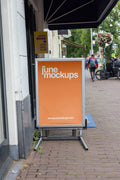 Amsterdam street A-board poster mockup in DIN format placed on a brick sidewalk in front of a local shop with realistic city background, pedestrians, bicycles and storefront details.