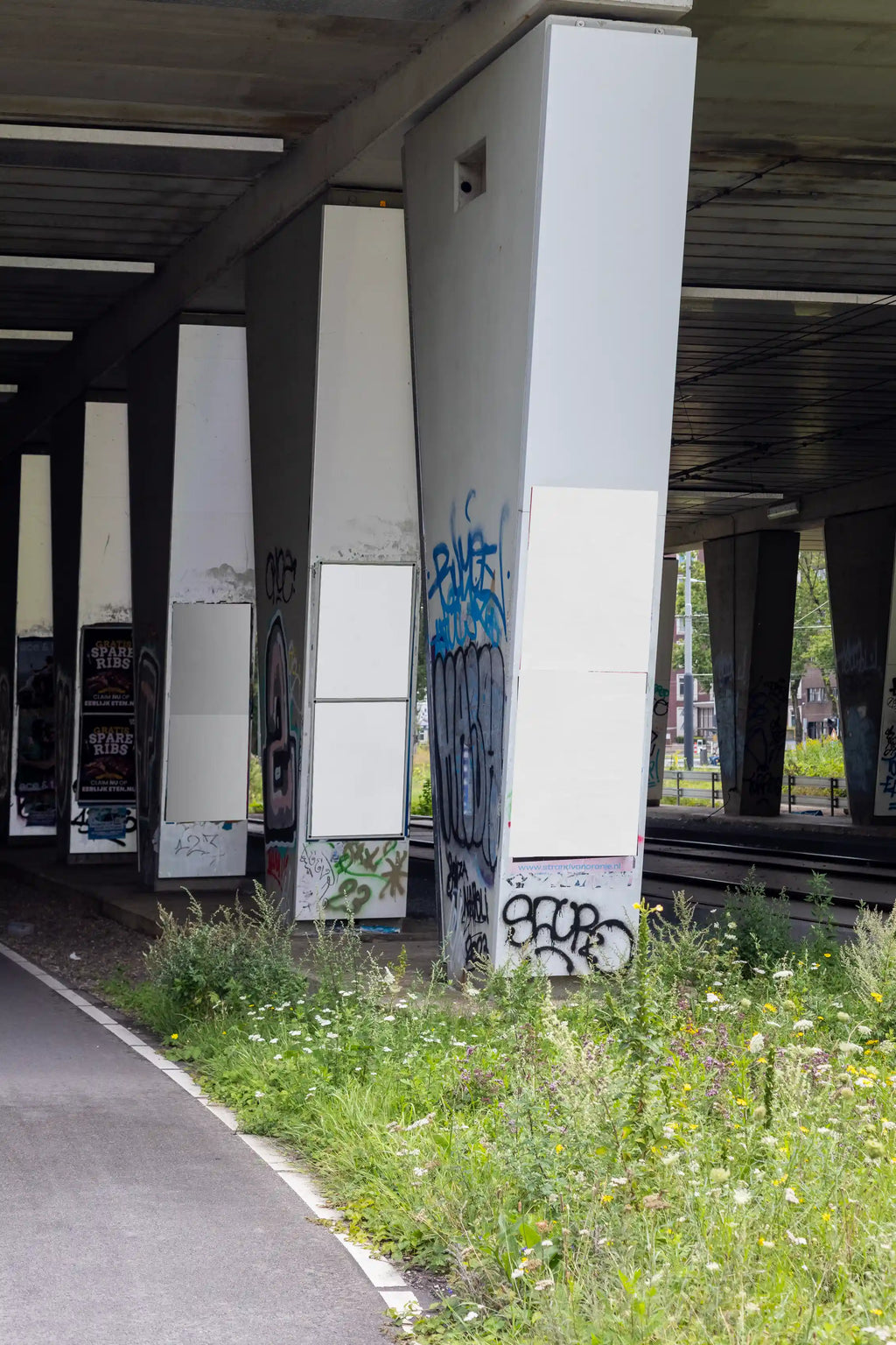 Blank background Amsterdam urban poster mockup under a concrete bridge featuring multiple DIN-format poster placements on large concrete columns with graffiti and natural greenery.
