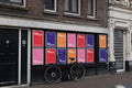 Amsterdam urban street poster mockups on Muntplein corner facade with multiple vibrant posters and a parked bike.