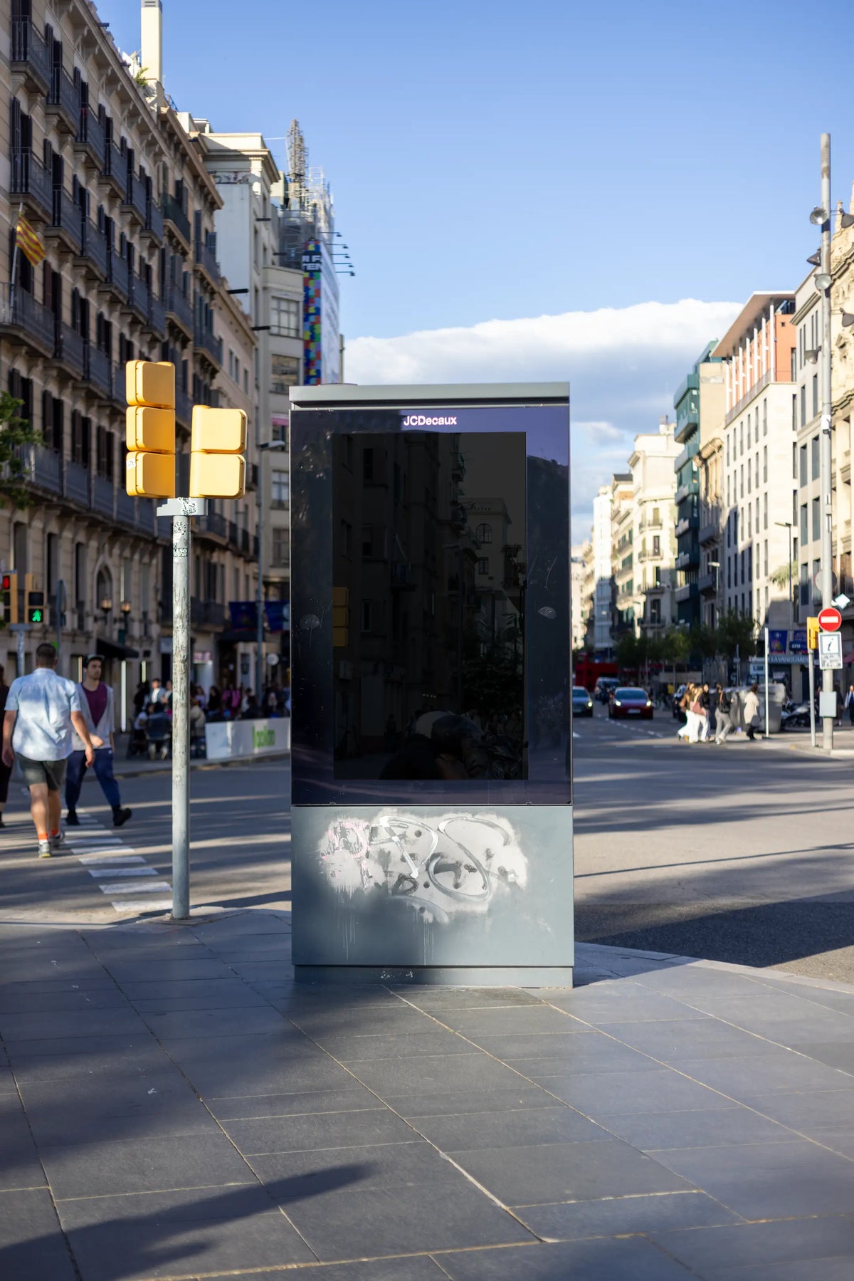 Black blank 9:16 advertising display in central Barcelona, featuring a reflective dark portrait panel inside a freestanding JCDecaux city light structure. Historic European architecture, yellow traffic signals, passing pedestrians and graffiti on the metal base create a realistic setting for a blank outdoor advertising mockup.
