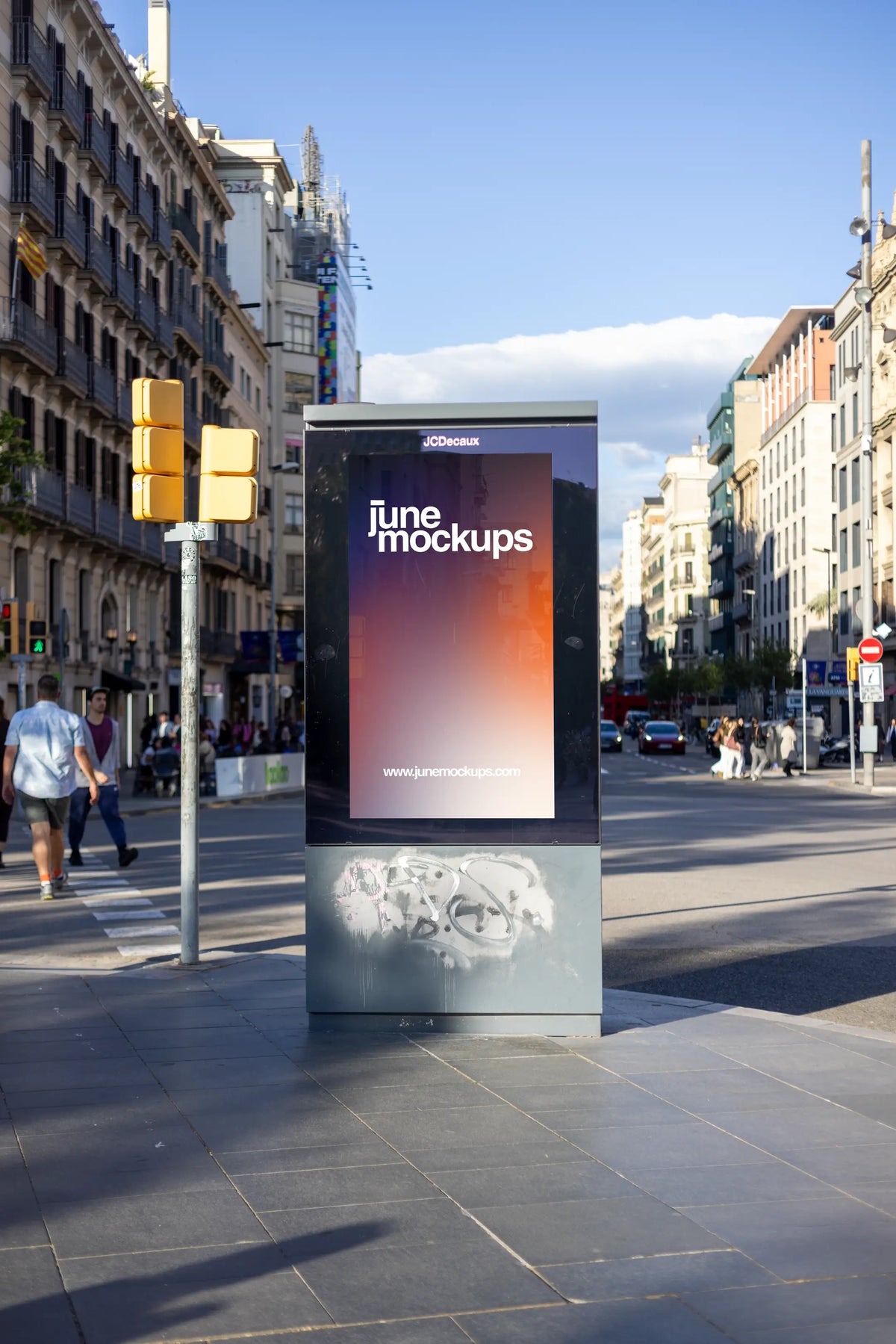 Realistic 9:16 city light advertising mockup photographed at a busy intersection in Barcelona, Spain. A freestanding JCDecaux poster display features a JuneMockups design, surrounded by historic buildings, balconies, yellow traffic lights and pedestrians. The graffiti-covered base adds authentic street character to this urban DOOH-style PSD presentation.