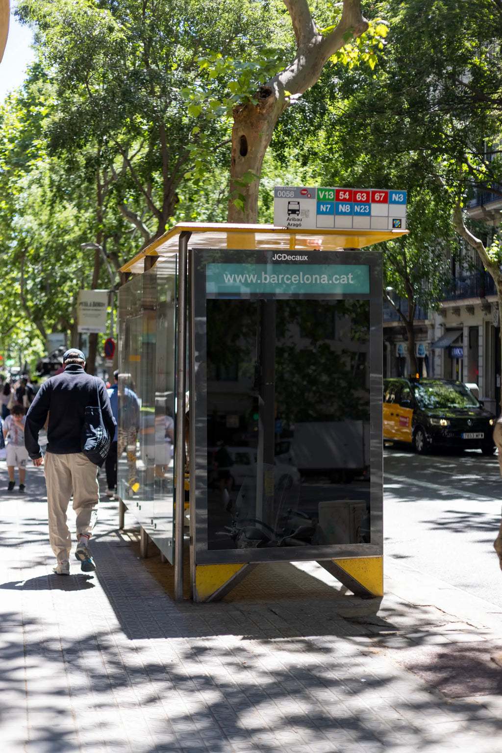 Empty black reflective JCDecaux city light poster display at the Aribau–Aragó bus stop in Barcelona's Eixample neighborhood. The tall vertical blank advertising panel sits inside a glass bus shelter with a yellow metal roof, visible public transport route numbers and a turquoise Barcelona information strip above the empty poster area. Large leafy plane trees frame the photograph, casting distinct patterns of sunlight and shade across the textured pavement. A pedestrian wearing a dark jacket walks beside the