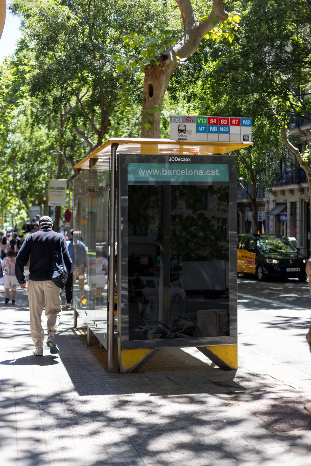 Empty black reflective JCDecaux city light poster display at the Aribau–Aragó bus stop in Barcelona's Eixample neighborhood. The tall vertical blank advertising panel sits inside a glass bus shelter with a yellow metal roof, visible public transport route numbers and a turquoise Barcelona information strip above the empty poster area. Large leafy plane trees frame the photograph, casting distinct patterns of sunlight and shade across the textured pavement. A pedestrian wearing a dark jacket walks beside the