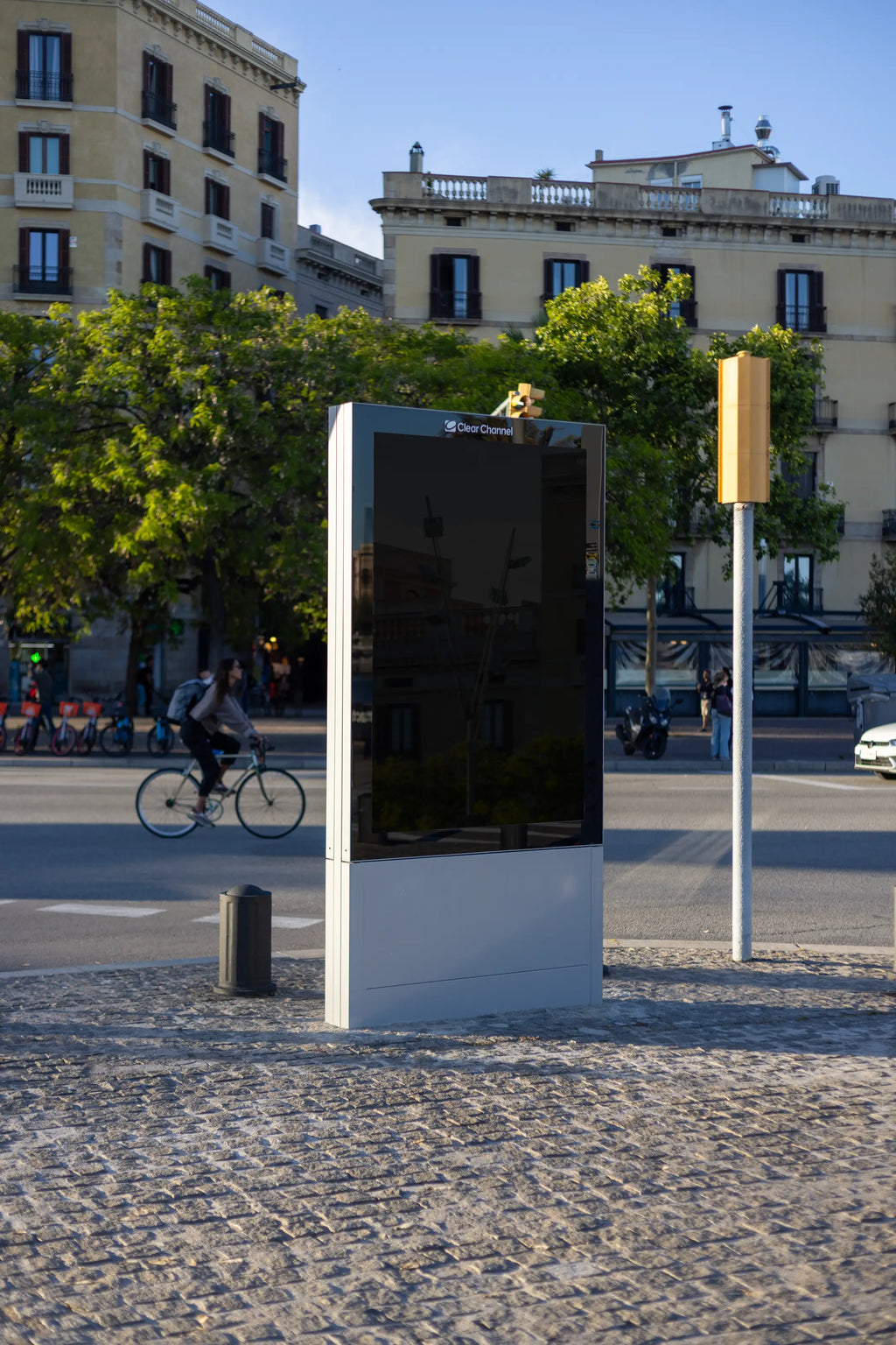 Black blank freestanding Clear Channel advertising display photographed on a sunlit cobblestone street in Barcelona, Spain. The tall vertical advertising panel features an empty dark reflective surface surrounded by a silver metal frame and a solid rectangular base. The structure is viewed from a slight side angle, emphasizing its realistic proportions and three-dimensional appearance. A cyclist passes through the street behind the display, while traditional Barcelona residential architecture, historic ston
