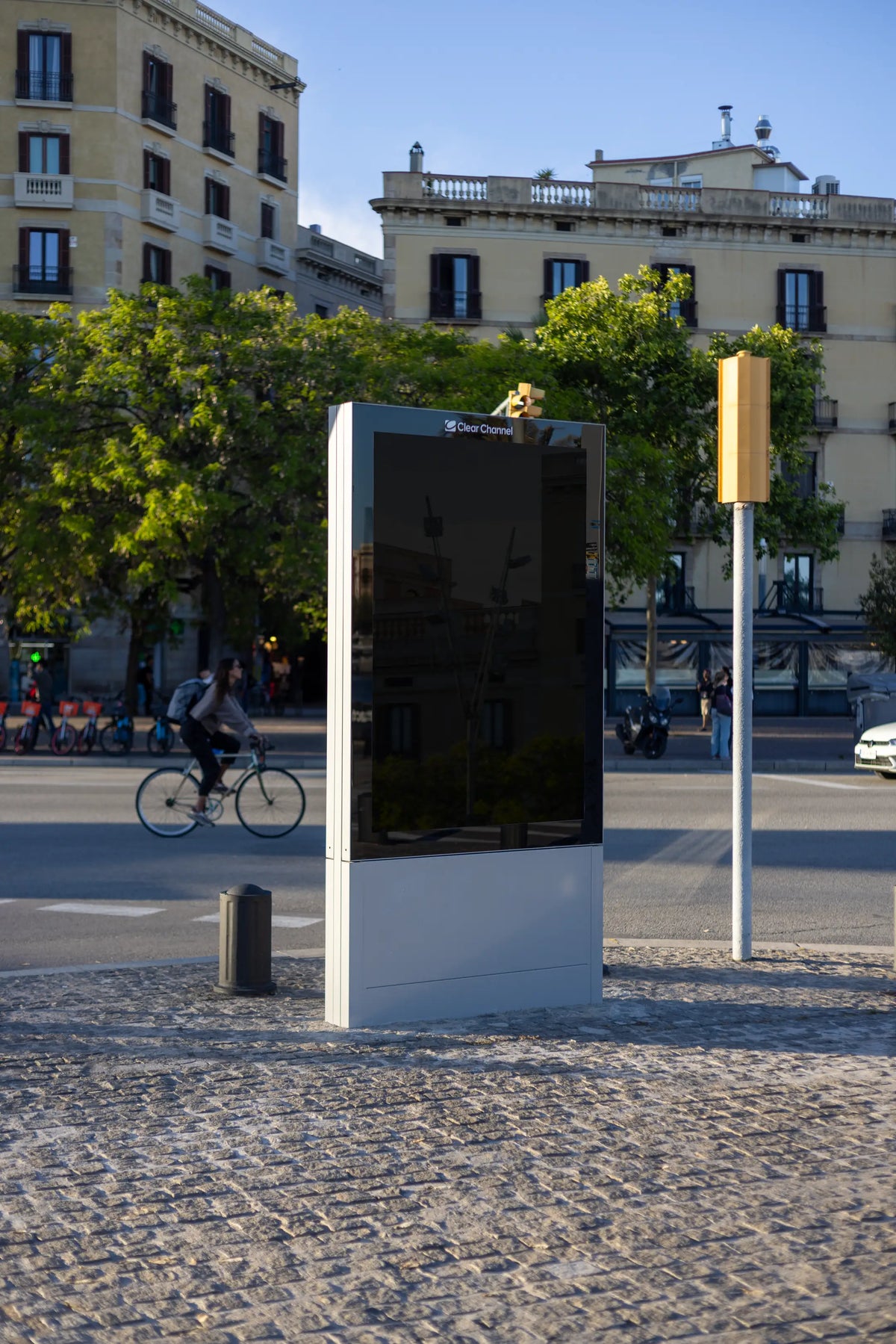 Black blank freestanding Clear Channel advertising display photographed on a sunlit cobblestone street in Barcelona, Spain. The tall vertical advertising panel features an empty dark reflective surface surrounded by a silver metal frame and a solid rectangular base. The structure is viewed from a slight side angle, emphasizing its realistic proportions and three-dimensional appearance. A cyclist passes through the street behind the display, while traditional Barcelona residential architecture, historic ston