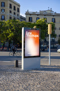 Freestanding Clear Channel city light poster mockup captured in a historic Barcelona street environment with warm afternoon sunlight. A modern outdoor advertising display with a silver metal frame, dark reflective border and large vertical poster area features a vibrant JuneMockups design. The display is photographed from a slightly angled perspective, creating realistic depth and visible side panels. Behind the advertising structure, a cyclist rides past traditional cream-colored residential buildings with
