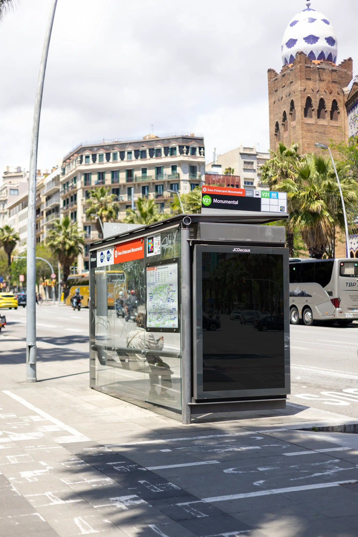 Black blank JCDecaux bus shelter advertising display photographed beside the historic La Monumental landmark on Carrer de la Marina in Barcelona's Eixample district, Spain. The empty vertical poster panel features a dark glossy reflective surface enclosed by a black metal advertising frame attached to a modern glass public transport shelter. Subtle reflections of passing vehicles, palm trees, pedestrians and surrounding buildings remain visible on the blank advertising surface, giving the scene an authentic