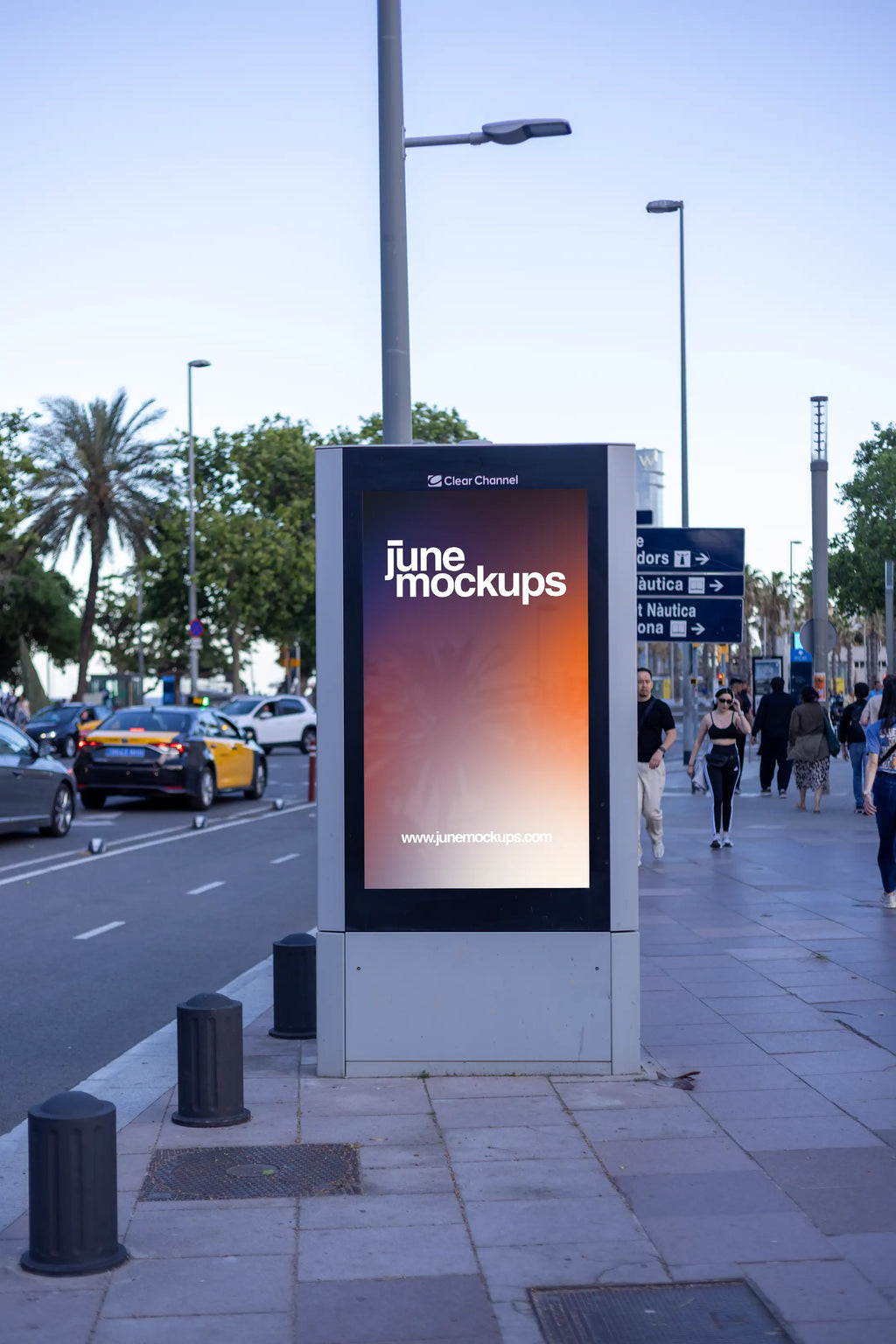 Photorealistic 9:16 DOOH-style city light advertising mockup photographed near Barcelona's marina and waterfront district. A modern freestanding Clear Channel outdoor advertising structure features a vertical portrait display with a colorful JuneMockups gradient design, surrounded by a dark reflective frame and silver-gray base. The advertising panel stands beside a busy Mediterranean avenue lined with tall palm trees, streetlights and wide pedestrian sidewalks. Blue road direction signs point toward Barcel