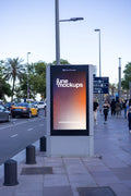 Photorealistic 9:16 DOOH-style city light advertising mockup photographed near Barcelona's marina and waterfront district. A modern freestanding Clear Channel outdoor advertising structure features a vertical portrait display with a colorful JuneMockups gradient design, surrounded by a dark reflective frame and silver-gray base. The advertising panel stands beside a busy Mediterranean avenue lined with tall palm trees, streetlights and wide pedestrian sidewalks. Blue road direction signs point toward Barcel
