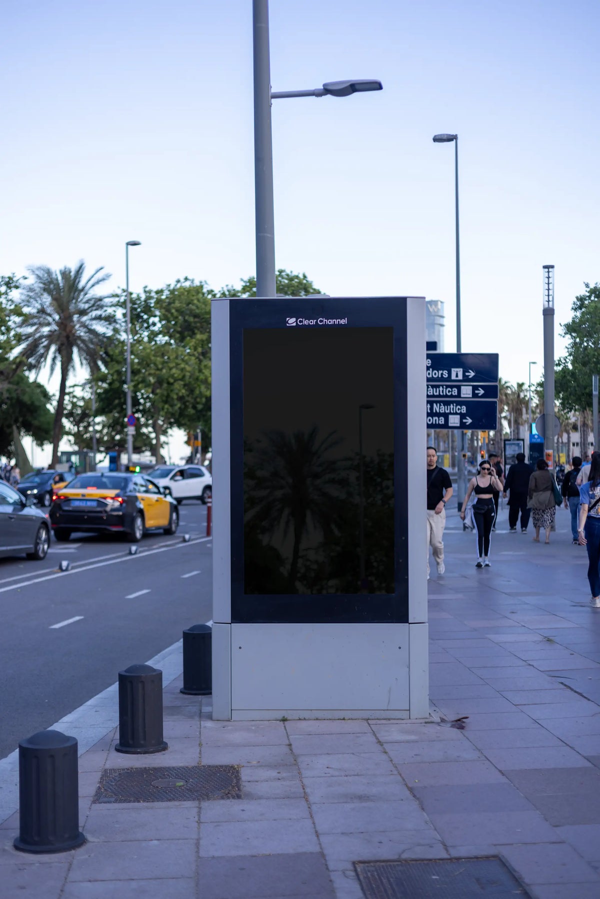 Empty black reflective 9:16 portrait display photographed inside a freestanding Clear Channel city light advertising structure near the Barcelona marina. The vertical blank panel is surrounded by a dark frame and a rectangular silver-gray metal base, with subtle reflections of the surrounding street environment visible on the surface. Mediterranean palm trees, nautical wayfinding signs, streetlights, pedestrians and Barcelona's distinctive black-and-yellow taxis create a realistic urban waterfront backdrop.