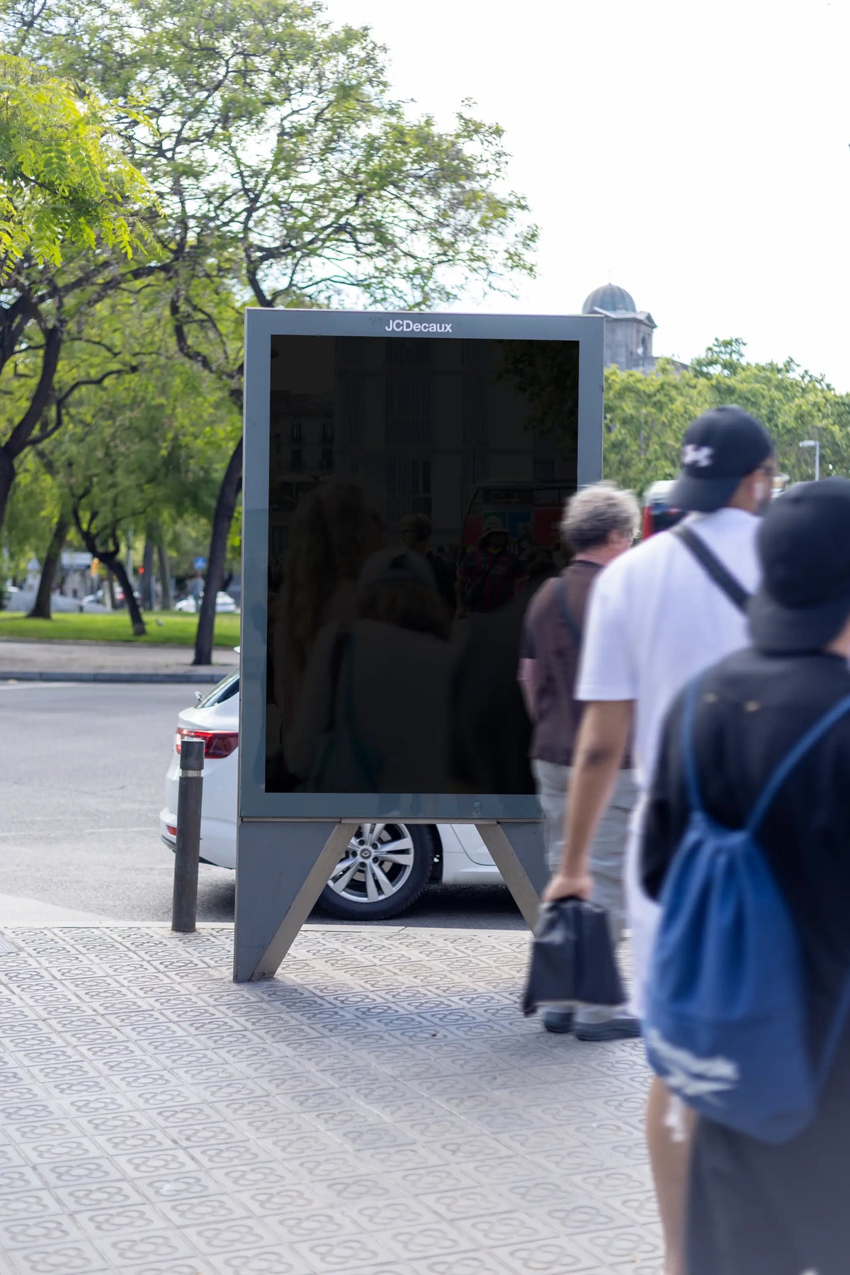 Black blank freestanding JCDecaux advertising display photographed on a patterned pedestrian sidewalk beside a green urban space in Barcelona, Spain. The empty reflective portrait poster panel is enclosed by a gray metal frame supported by an elevated base with angular legs. Several pedestrians wearing casual streetwear walk close to the camera, partially covering the right side of the blank advertising surface and creating natural foreground depth. A white parked car, leafy trees, distant pedestrians and a