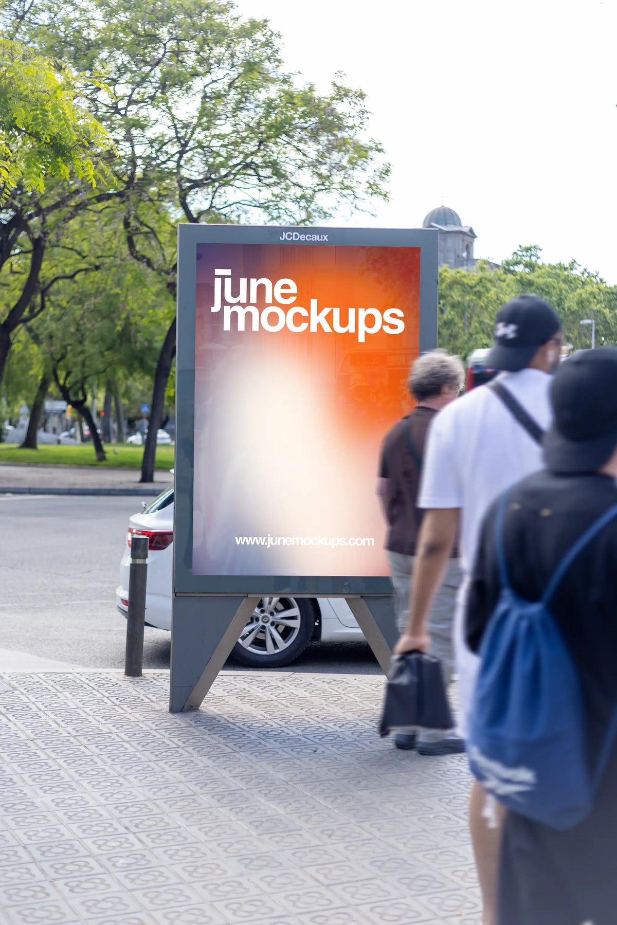 Photorealistic freestanding JCDecaux city light poster mockup captured beside a leafy urban green space in Barcelona, Spain. A large vertical outdoor advertising display with a gray metal frame and open triangular support base features a colorful JuneMockups gradient design. Several pedestrians pass directly in front of the advertising structure, partially obscuring the right side of the poster and adding spontaneous movement to the composition. Mature trees, a parked white car, decorative Barcelona pavemen