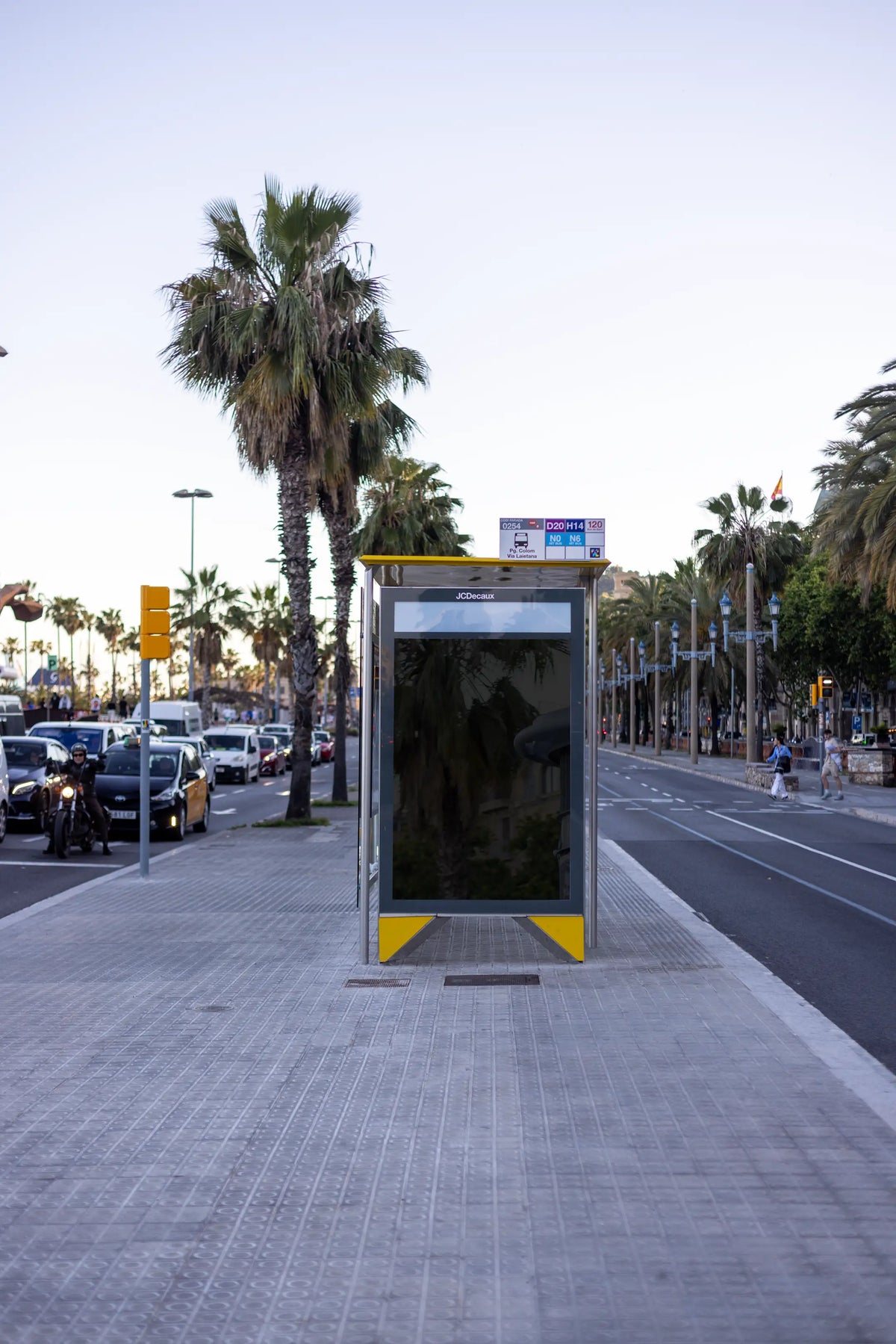Empty black reflective bus shelter advertising panel photographed on Passeig de Colom in Barcelona, Spain, near Via Laietana and the city's Mediterranean waterfront. The blank vertical poster surface is positioned inside a JCDecaux public transport shelter featuring a yellow roof structure, glass panels and a bus stop information display above the advertising frame. Large palm trees rise beside the pavement, with Barcelona taxis, motorcycles, moving vehicles and distant pedestrians along the avenue. The dar