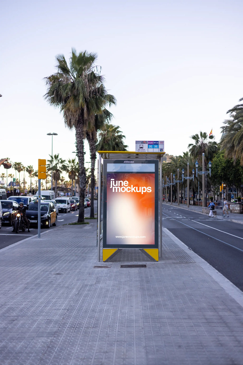Barcelona bus shelter city light poster mockup photographed on Passeig de Colom near Via Laietana, featuring a vertical JCDecaux city light advertising panel with a colorful JuneMockups design. Tall palm trees, a yellow bus shelter structure, Mediterranean boulevard, moving traffic and Barcelona taxis create an authentic waterfront setting for outdoor advertising, travel branding and OOH poster presentations.