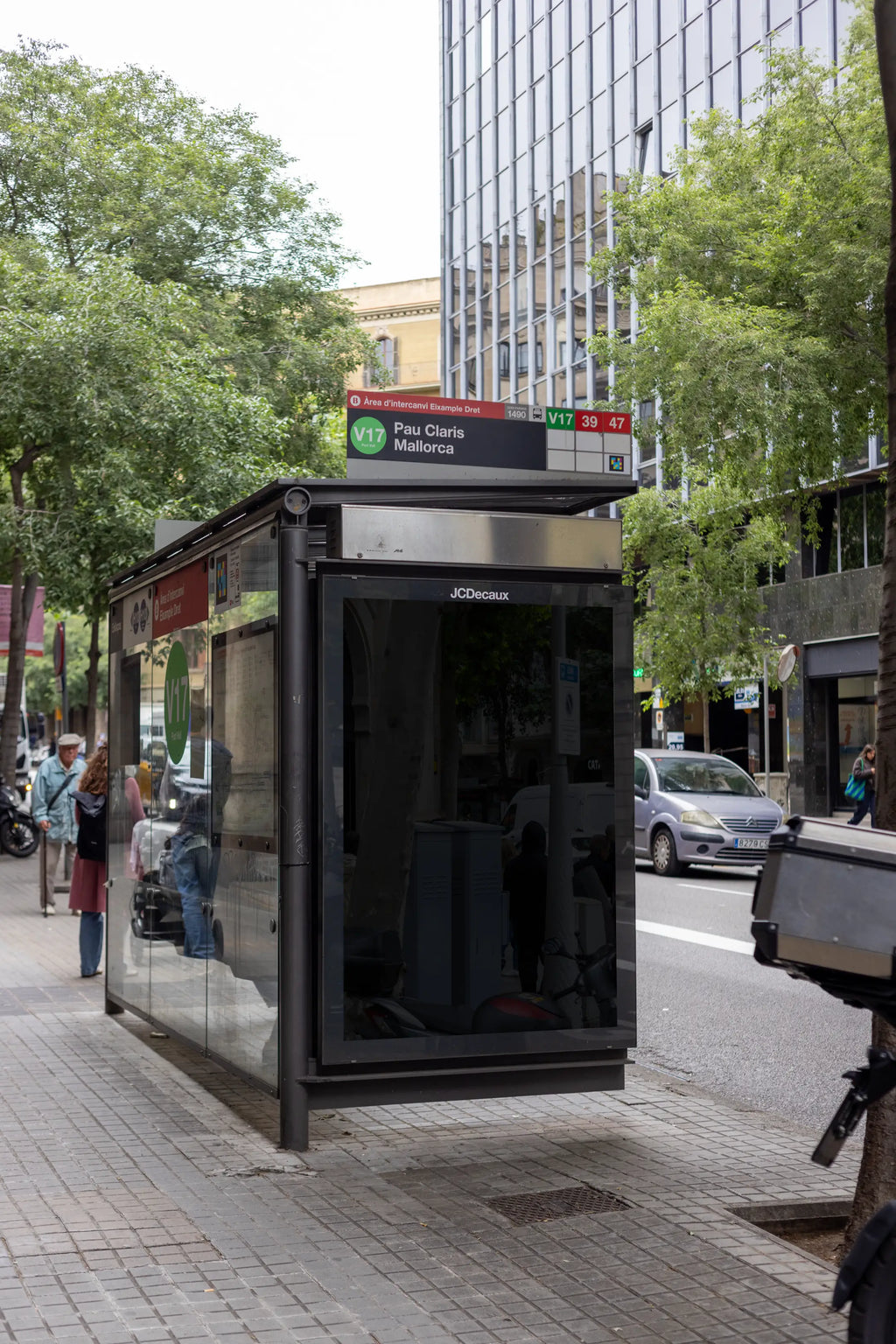 Black blank vertical advertising panel inside a JCDecaux bus shelter at Pau Claris–Mallorca in the Eixample neighborhood of Barcelona, Spain. The empty dark reflective poster surface is enclosed by a black metal frame attached to a modern glass public transport shelter with a silver roof, route signage and transparent side panels. The slightly angled perspective reveals reflections in the glass, a contemporary office building with repetitive vertical windows and mature leafy trees lining the avenue. A silve