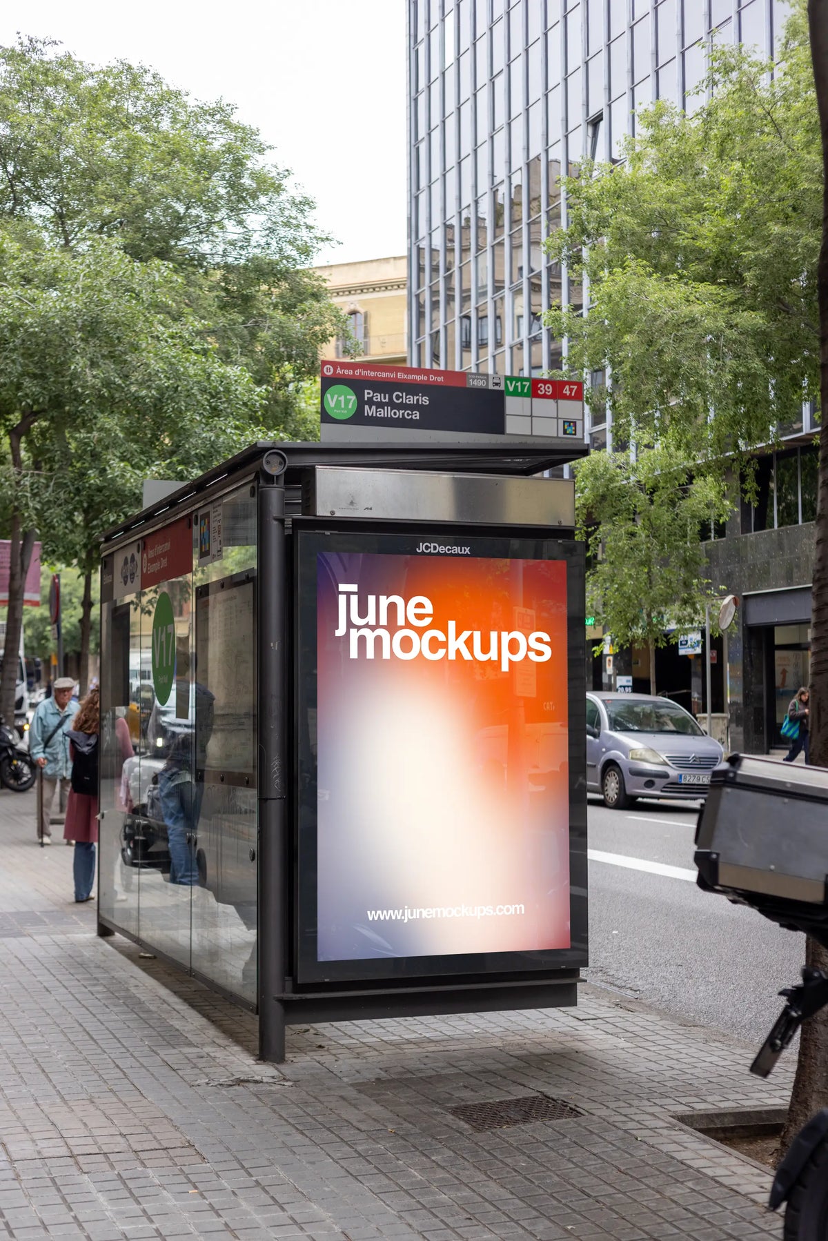 Photorealistic bus shelter poster mockup photographed at the Pau Claris–Mallorca public transport stop in Barcelona's Eixample district, Spain. A vertical JCDecaux advertising panel featuring a colorful JuneMockups design is integrated into a modern glass bus shelter with a brushed metal roof and visible route information signage. The image is captured from a slightly angled street-level perspective, revealing the transparent side panels and natural reflections of the surrounding environment. A contemporary