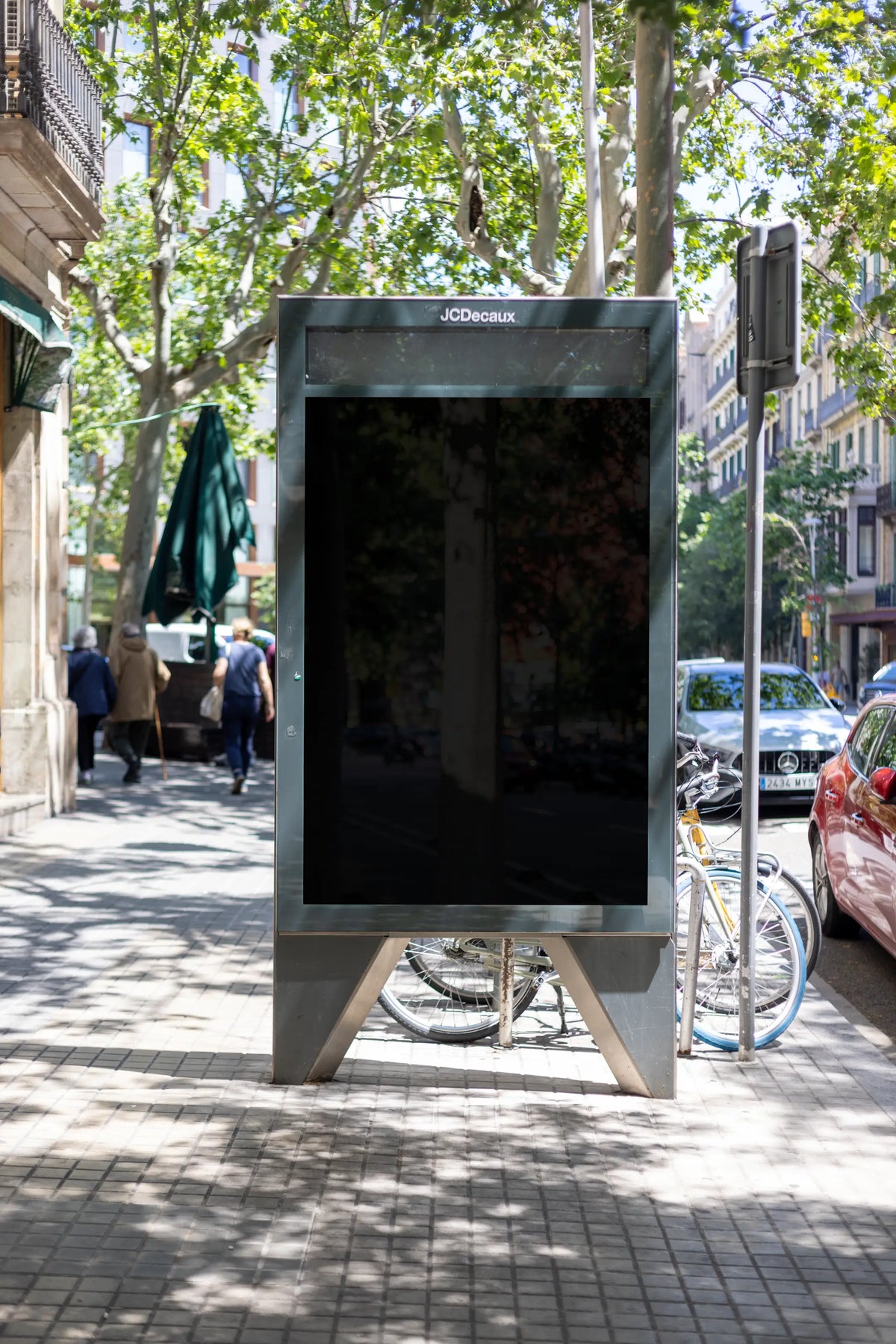 Black blank JCDecaux freestanding city light advertising display photographed on a bright tree-lined sidewalk in Barcelona, Spain. The empty dark reflective poster panel is framed by a rectangular gray metal structure with a raised base and angular support legs. Above the display, large green plane trees extend across the street, casting scattered sunlight and natural leaf shadows onto the patterned pavement. Parked bicycles, scooters, cars, folded café umbrellas and pedestrians appear along the sidewalk, w