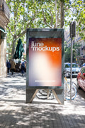 Realistic Barcelona avenue poster mockup featuring a large freestanding JCDecaux city light advertising display with a colorful JuneMockups gradient design. The portrait poster panel is enclosed by a dark gray metal frame with angular support legs and positioned beneath the green canopy of mature plane trees along a sunlit pedestrian sidewalk. Traditional Barcelona residential buildings with wrought-iron balconies and classical architectural details line the street, while bicycles, parked cars, pedestrians 
