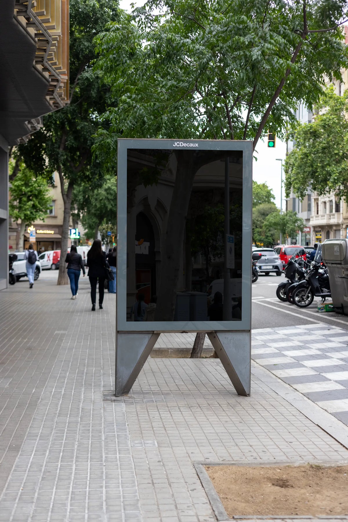 Blank black JCDecaux city light advertising display standing beside a pedestrian crossing on a tree-lined street in Barcelona, Spain. The freestanding metal structure features a large empty reflective poster panel enclosed by a gray frame, with a distinctive raised base and visible support legs beneath the advertising surface. Green leafy trees, traditional residential buildings with tall windows and balconies, passing cyclists and pedestrians provide a natural urban background. A yellow traffic signal, par