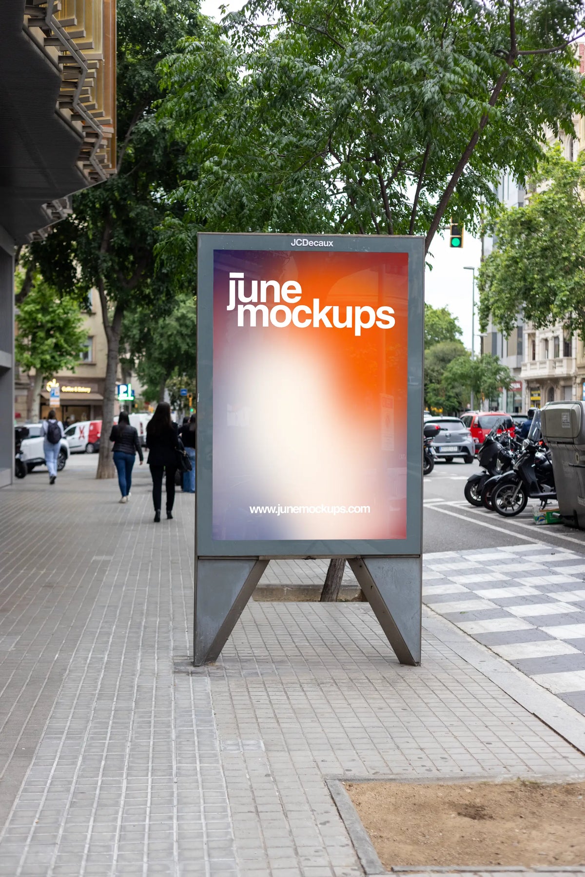 Realistic freestanding JCDecaux street poster mockup photographed along a leafy neighborhood sidewalk in Barcelona, Spain. A vertical outdoor advertising display with a gray metal frame and elevated open-base construction features a bright JuneMockups gradient poster. Dense green tree foliage provides a natural background, while historic cream-colored residential façades, wrought-iron balconies and pedestrians walking along the pavement establish a recognizable Barcelona street atmosphere. A pedestrian cros