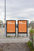 Two orange city light poster mockups in DIN format placed side by side near the water at Gannet / Holzpark Klybeck in Basel. The scene includes gravel, grass, cloudy sky, urban infrastructure and a quiet outdoor setting, perfect for out-of-home advertising, branding and poster campaign presentations.