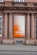 Vertical 1:2 banner mockup on the facade of the Historisches Museum Basel, Haus zum Kirschgarten. The large orange and white banner hangs between historic stone columns on a reddish sandstone building, with sidewalk and street curb visible below.