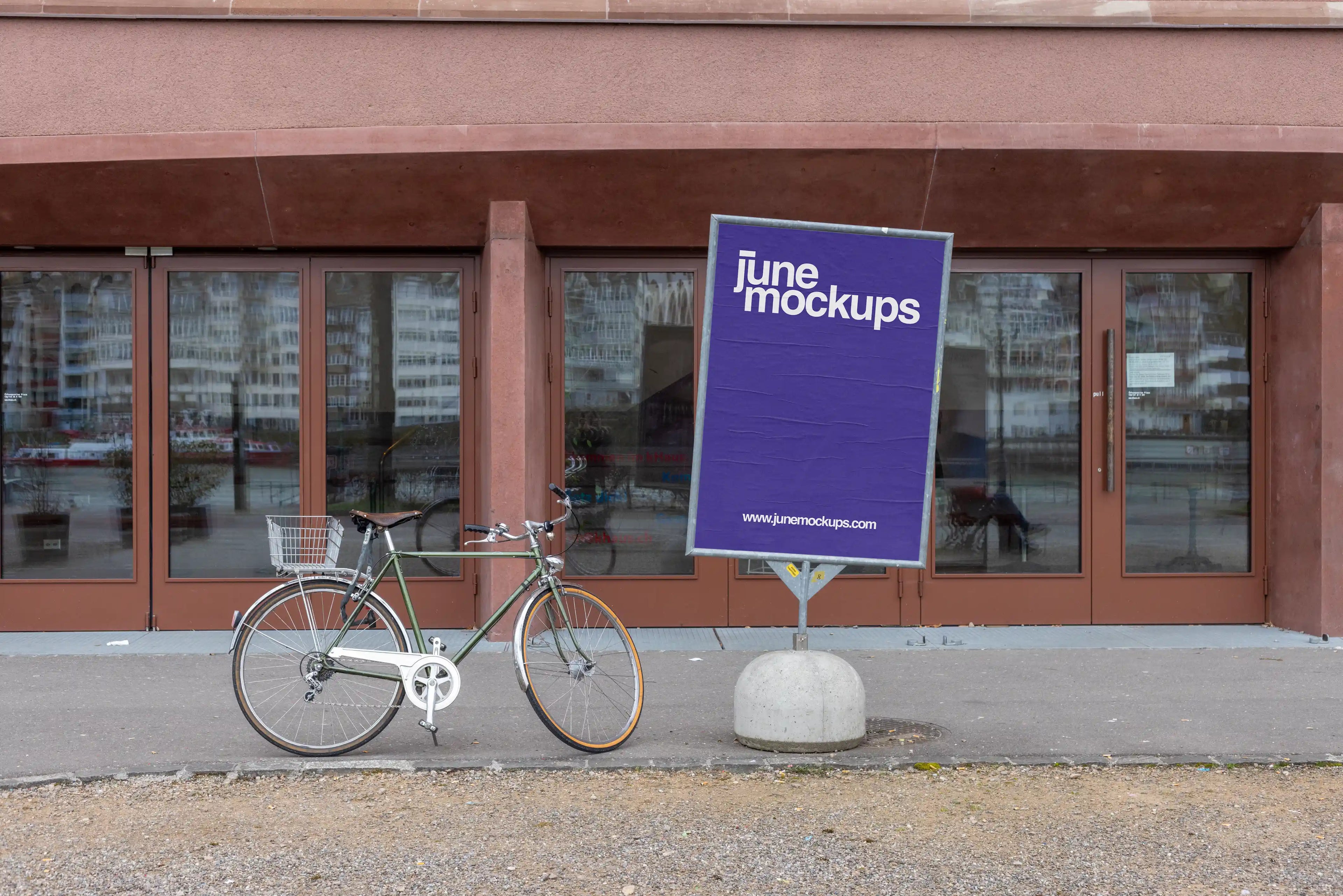 Purple DIN poster mockup on a freestanding metal poster display at Kaserne Basel near the Rhine riverside. A green bicycle stands next to the display, with glass doors, reflections, reddish-brown architecture, gravel ground and an urban cultural venue setting in the background.