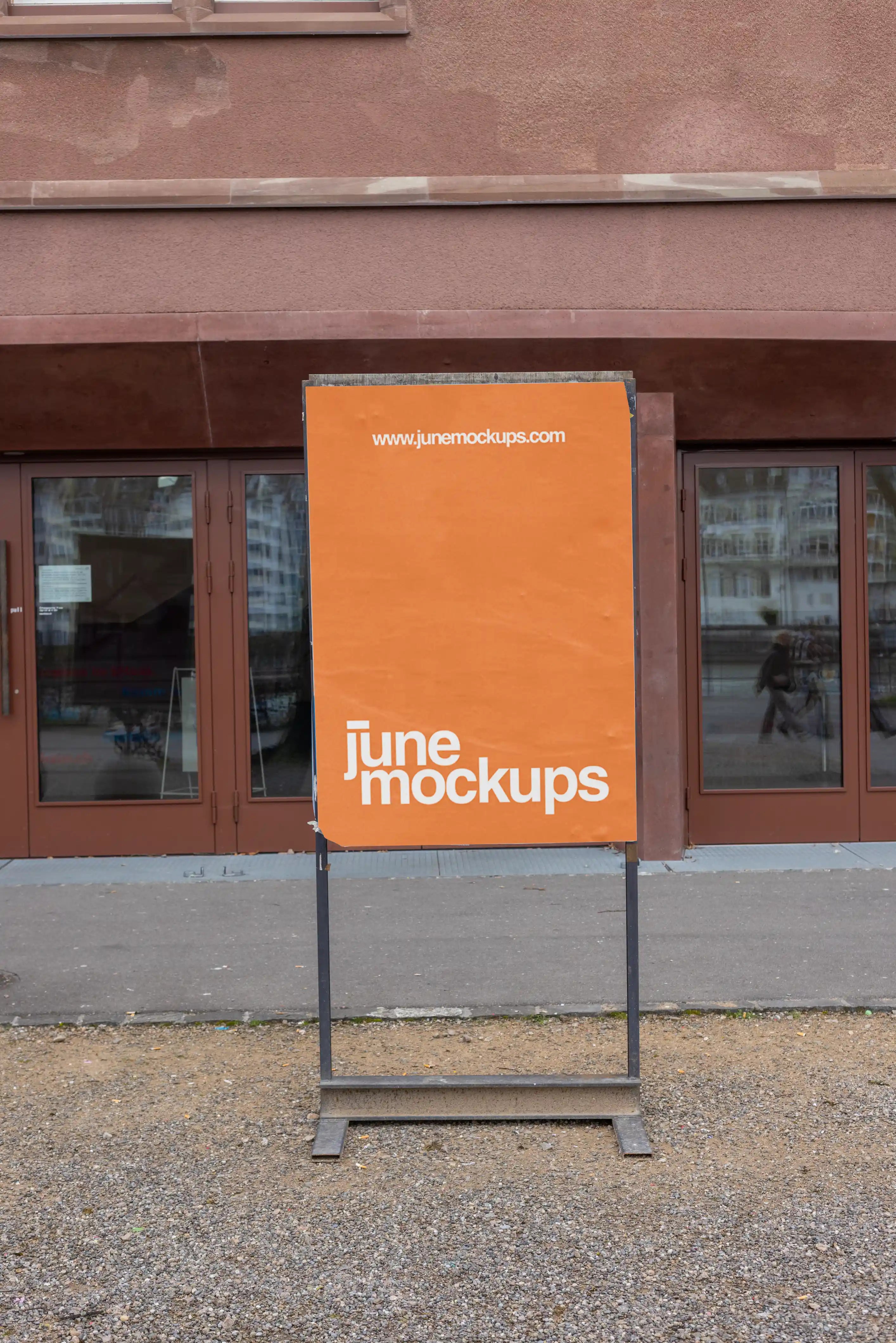 Orange vertical poster mockup on a freestanding outdoor sign display at Kaserne Basel on the Rhine side. The sign stands on a metal frame in front of reddish-brown glass doors, with gravel, pavement, subtle reflections and a clean public venue setting.