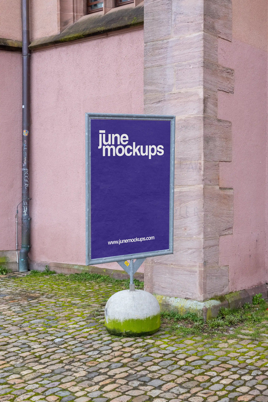 Single vertical DIN poster mockup at Kaserne Basel near Ausstellungsraum Klingental, placed on a freestanding metal display with a round concrete base. The scene shows a pink historic wall, stone corner, cobblestone pavement and moss details, suitable for poster design, event promotion and outdoor advertising previews.