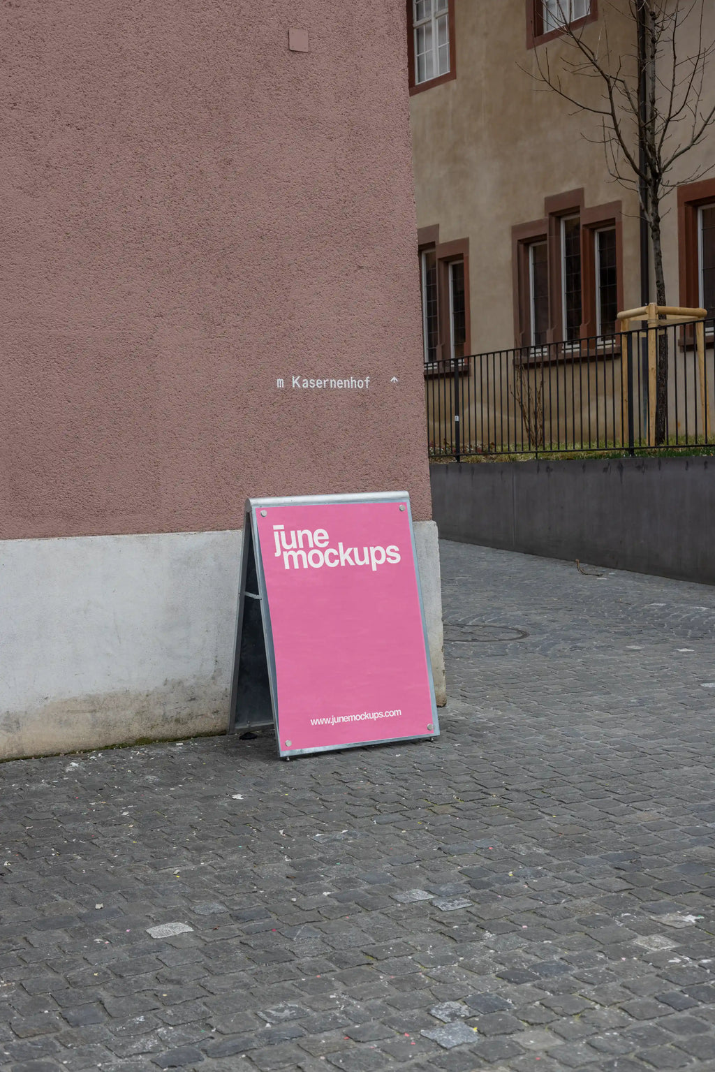 Pink DIN poster mockup attached to a freestanding magnetic board with visible metal corner magnets at Kasernenhof in Basel. The board leans on a cobblestone pathway beside a pink textured wall, with the word Kasernenhof visible in the background.