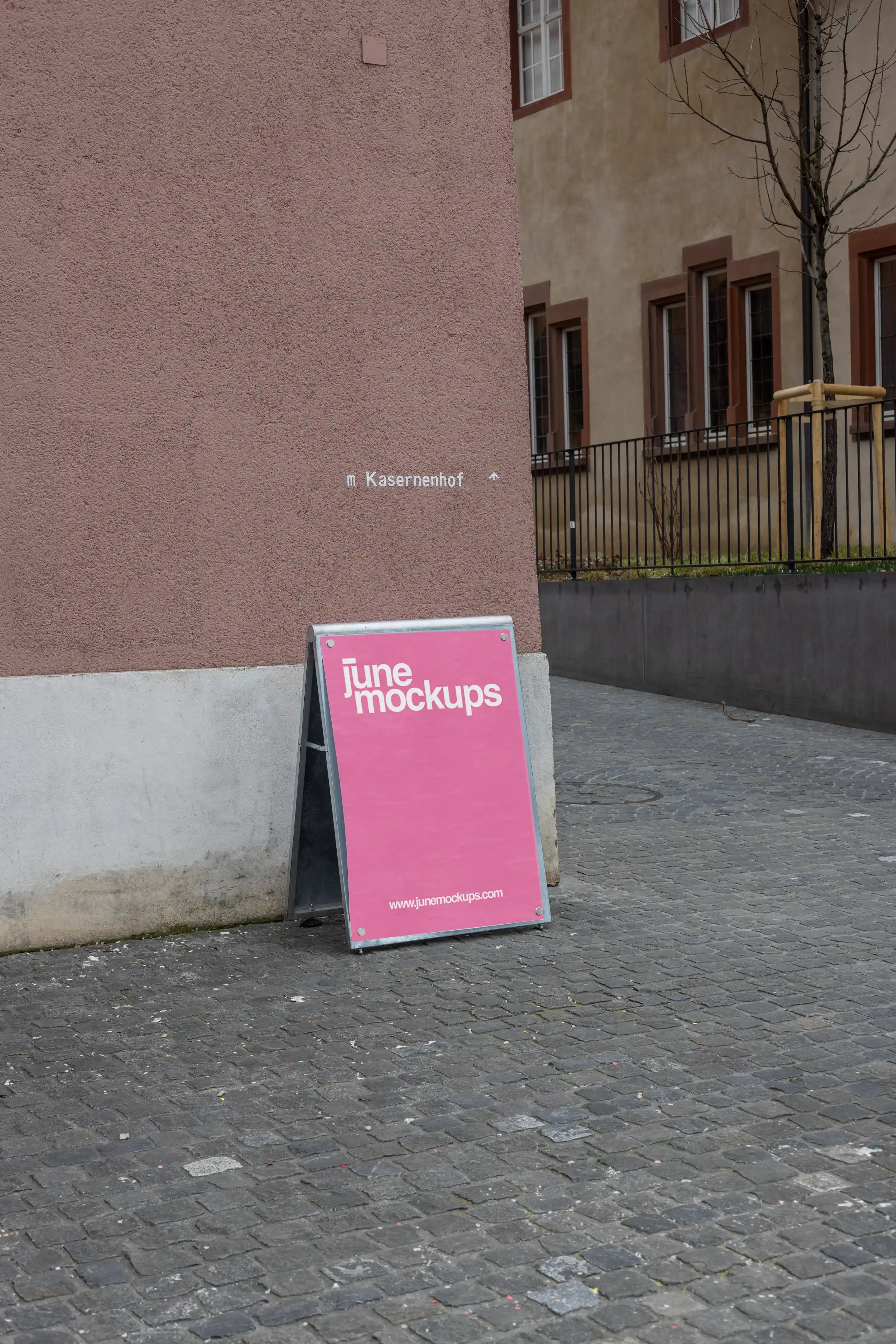 Pink DIN poster mockup attached to a freestanding magnetic board with visible metal corner magnets at Kasernenhof in Basel. The board leans on a cobblestone pathway beside a pink textured wall, with the word Kasernenhof visible in the background.