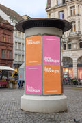 Cylindrical advertising column mockup at Basel Marktplatz with four vertical DIN poster placements in orange and pink. The scene is surrounded by historic buildings, cobblestone pavement, bicycles, market details and a lively city street atmosphere, ideal for Litfaßsäule and ad column poster previews.