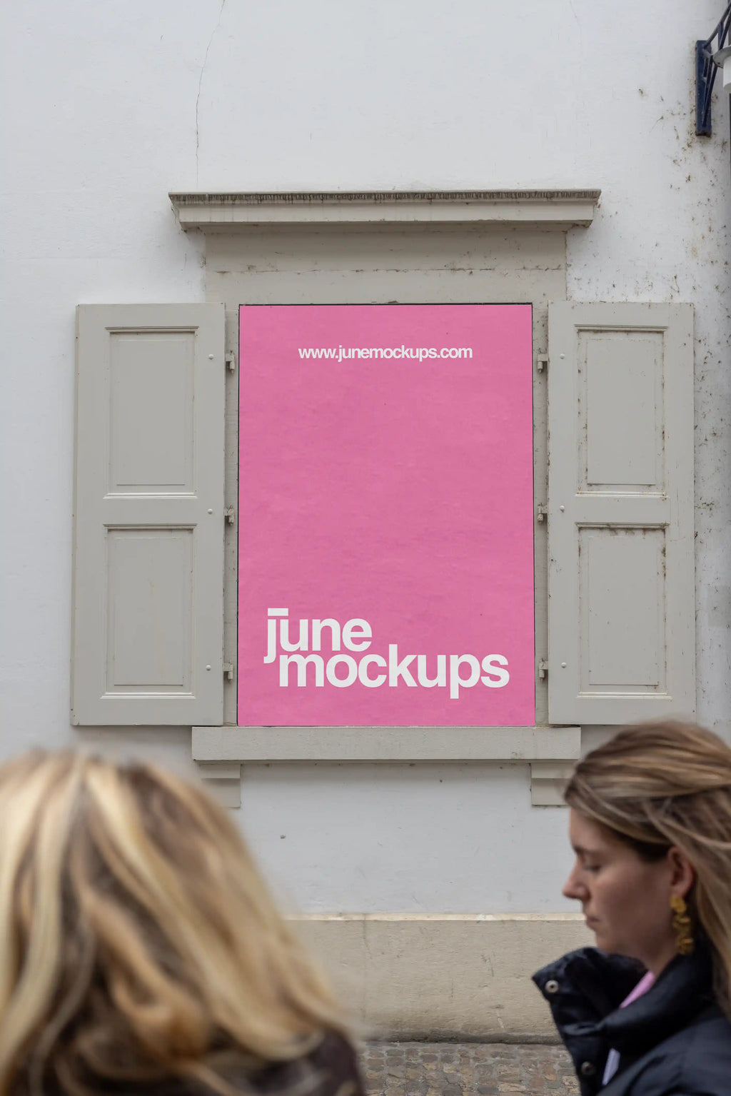 Pink DIN poster mockup placed in a window display at the Museum of Cultures Basel near Münsterplatz. Two blurred people pass in the foreground, while the poster sits between pale shutters on a white building facade, creating a realistic cultural street scene.