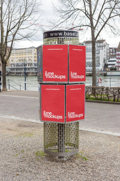 Four red DIN poster mockups on a modern cylindrical ad column near the Rhine riverside in Basel. The advertising column stands on gravel with trees, city buildings, water, sidewalk details and a calm urban waterfront atmosphere in the background.