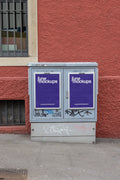 Two purple DIN poster mockups displayed in silver frames on a grey utility box in Basel. The utility box has graffiti and street marks, standing in front of a textured red wall with a barred window on the left, creating a raw urban poster mockup scene.