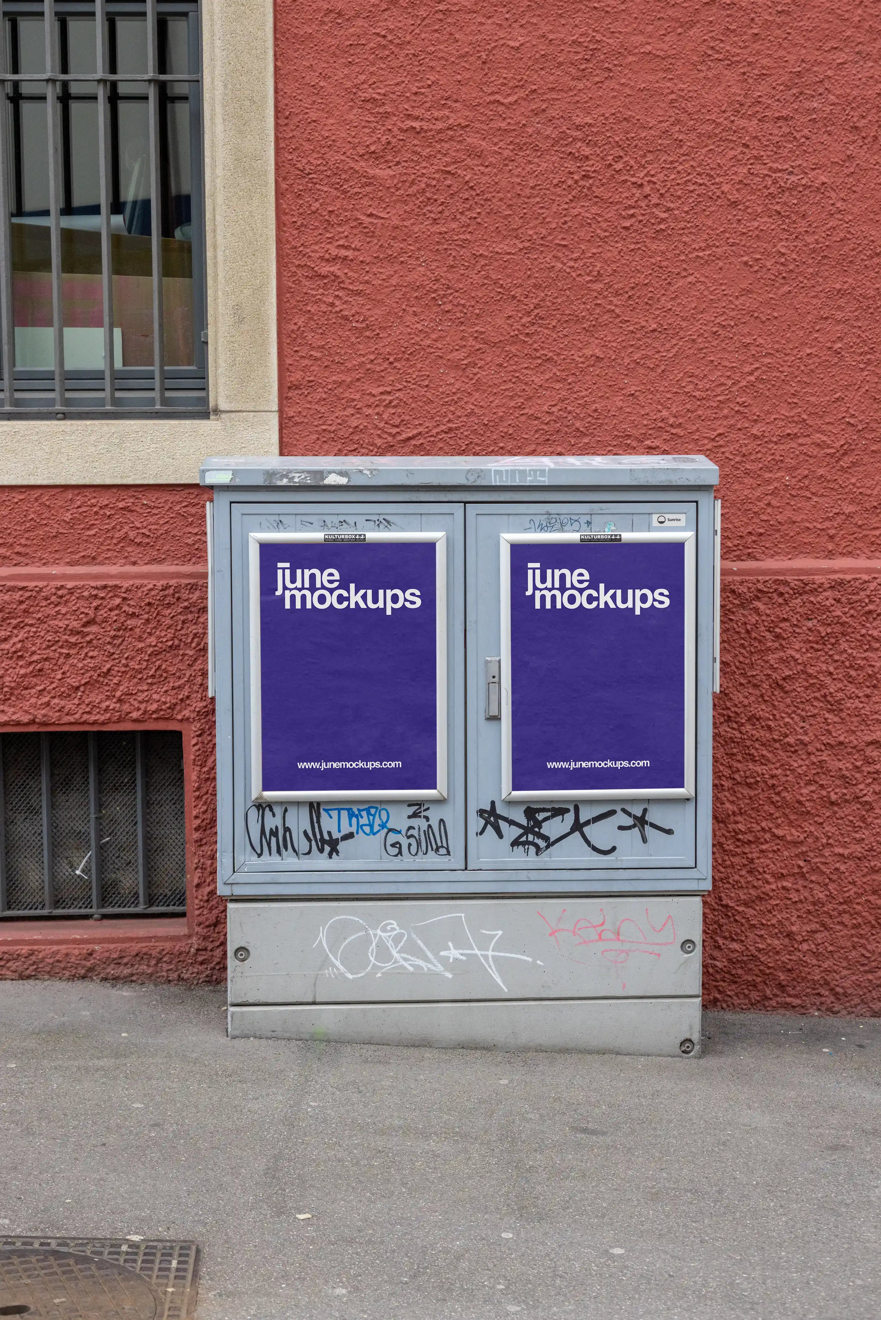 Two purple DIN poster mockups displayed in silver frames on a grey utility box in Basel. The utility box has graffiti and street marks, standing in front of a textured red wall with a barred window on the left, creating a raw urban poster mockup scene.