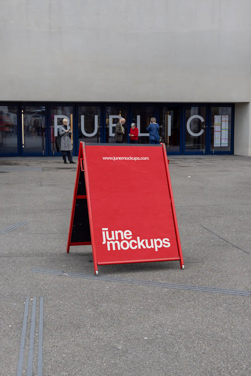 Red A-frame poster mockup placed outside a public entrance near Theater Basel. The sign stands on a grey pavement in front of large glass doors with the word public visible, while several people stand in the background near the entrance.