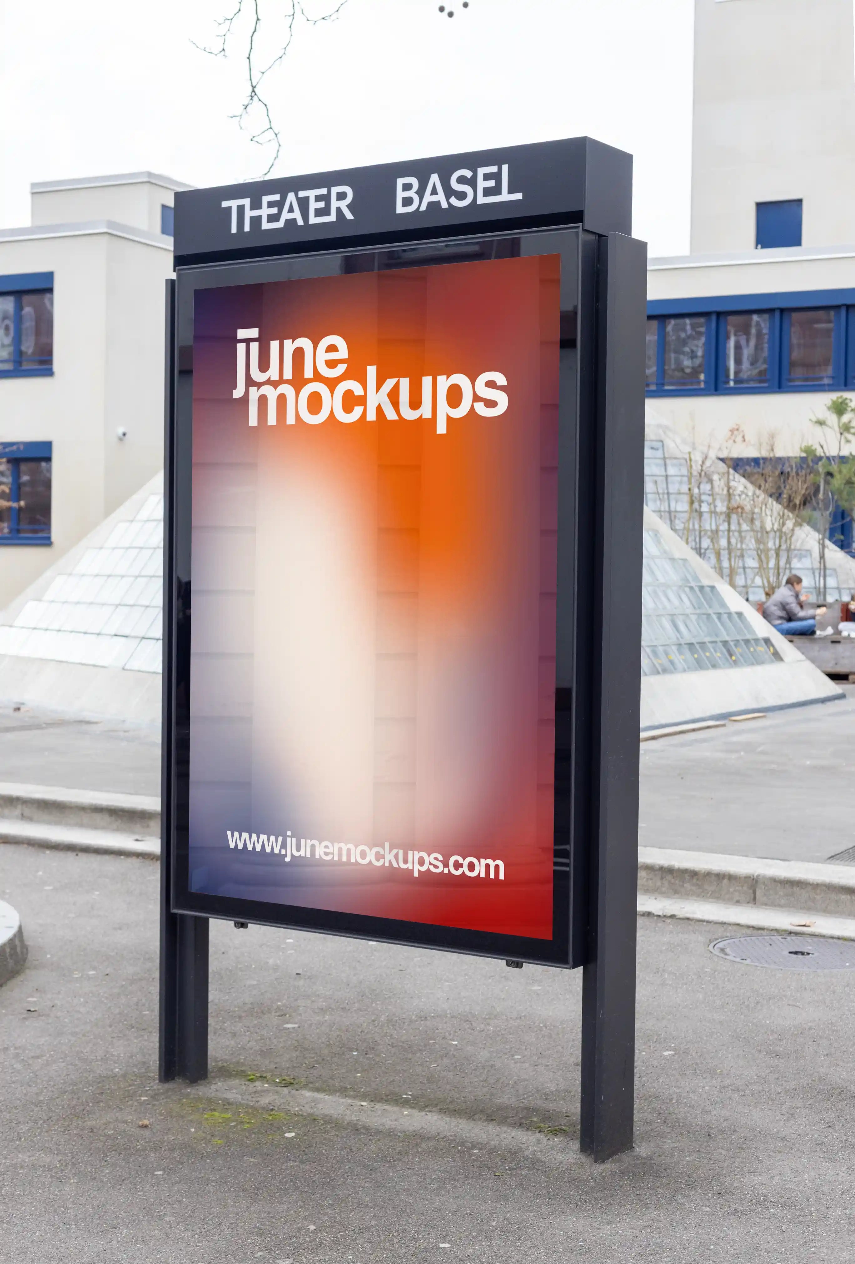 DIN format city light poster mockup in front of Theater Basel, placed inside a black outdoor advertising display. The scene shows the Theater Basel sign above the frame, modern architecture, glass pyramid structures, pavement and natural city light.