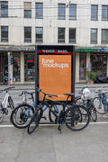 Orange DIN poster mockup inside a black city light display near Theater Basel. Several bicycles are parked in front of the display, with tram wires, storefronts, street details and urban architecture visible in the background.