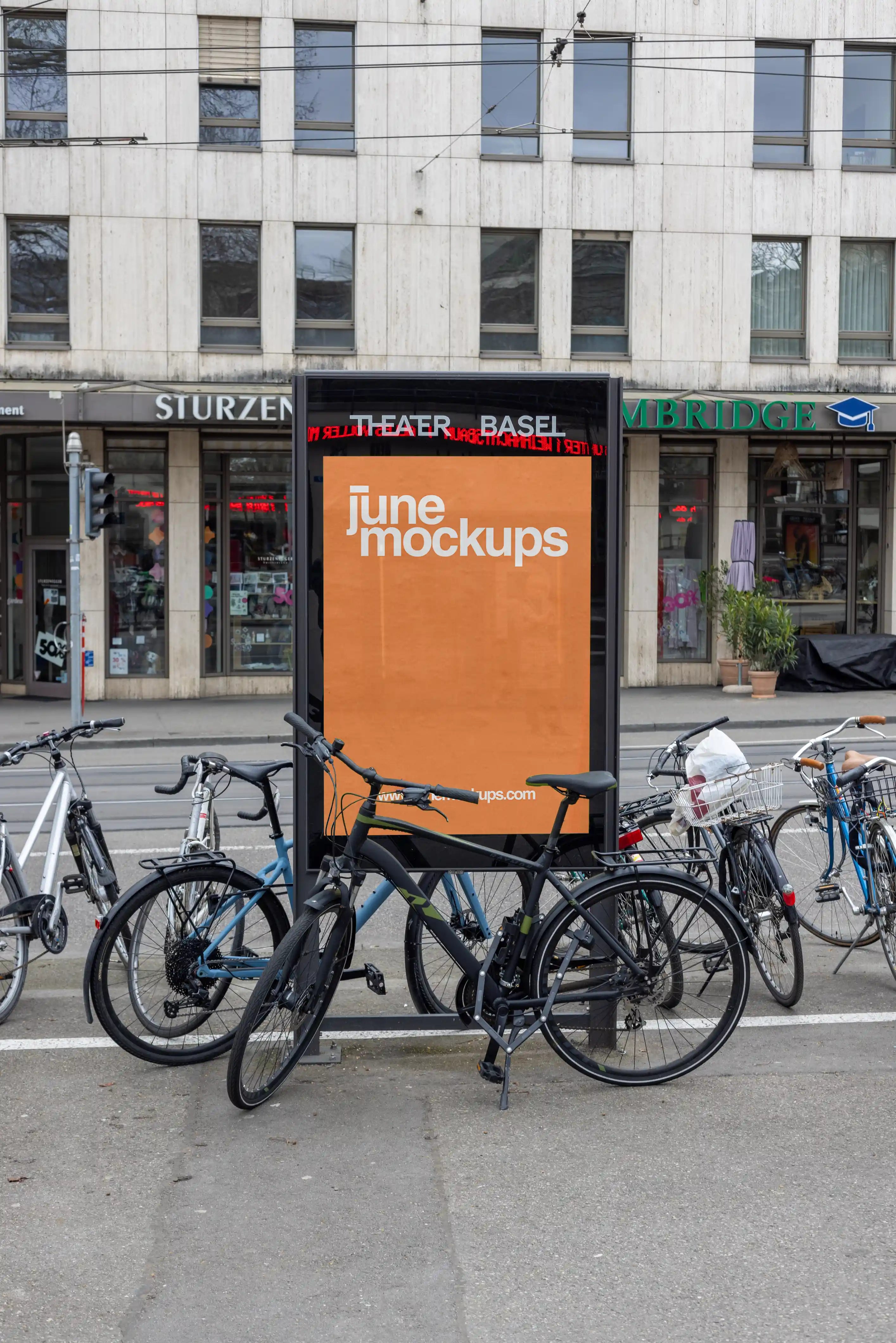 Orange DIN poster mockup inside a black city light display near Theater Basel. Several bicycles are parked in front of the display, with tram wires, storefronts, street details and urban architecture visible in the background.