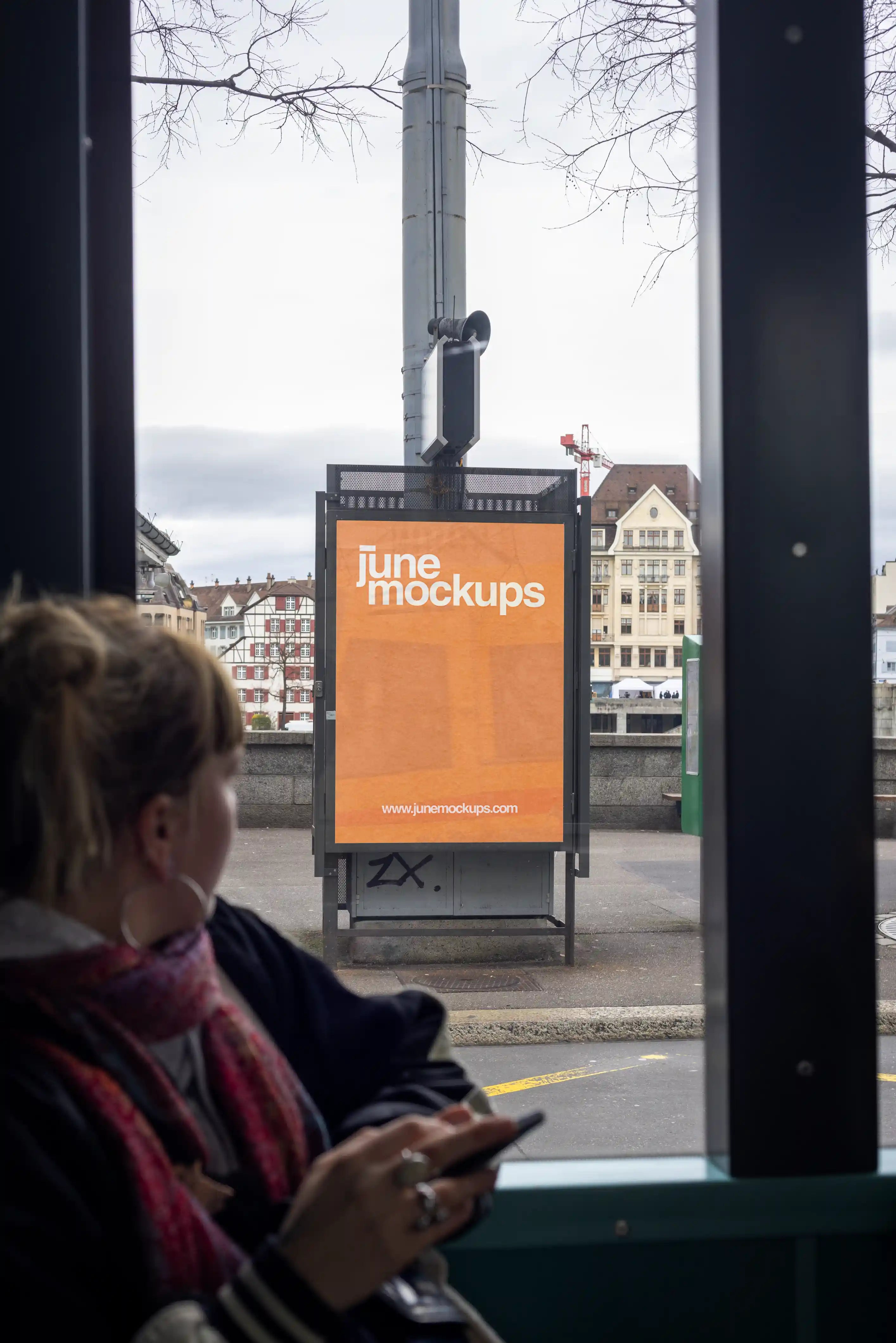 Orange DIN poster mockup seen from inside a tram in Basel, with the poster display outside near Mittlere Brücke. A seated passenger is blurred in the foreground, while the street, historic buildings, traffic pole and bridge area appear through the tram window.