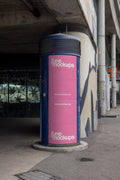 Pink two-part DIN poster mockup on a blue cylindrical advertising column in Basel under a concrete bridge structure. The scene includes urban shadows, worn surfaces, concrete architecture and street details, made for realistic Litfaßsäule, ad column and poster campaign presentations.