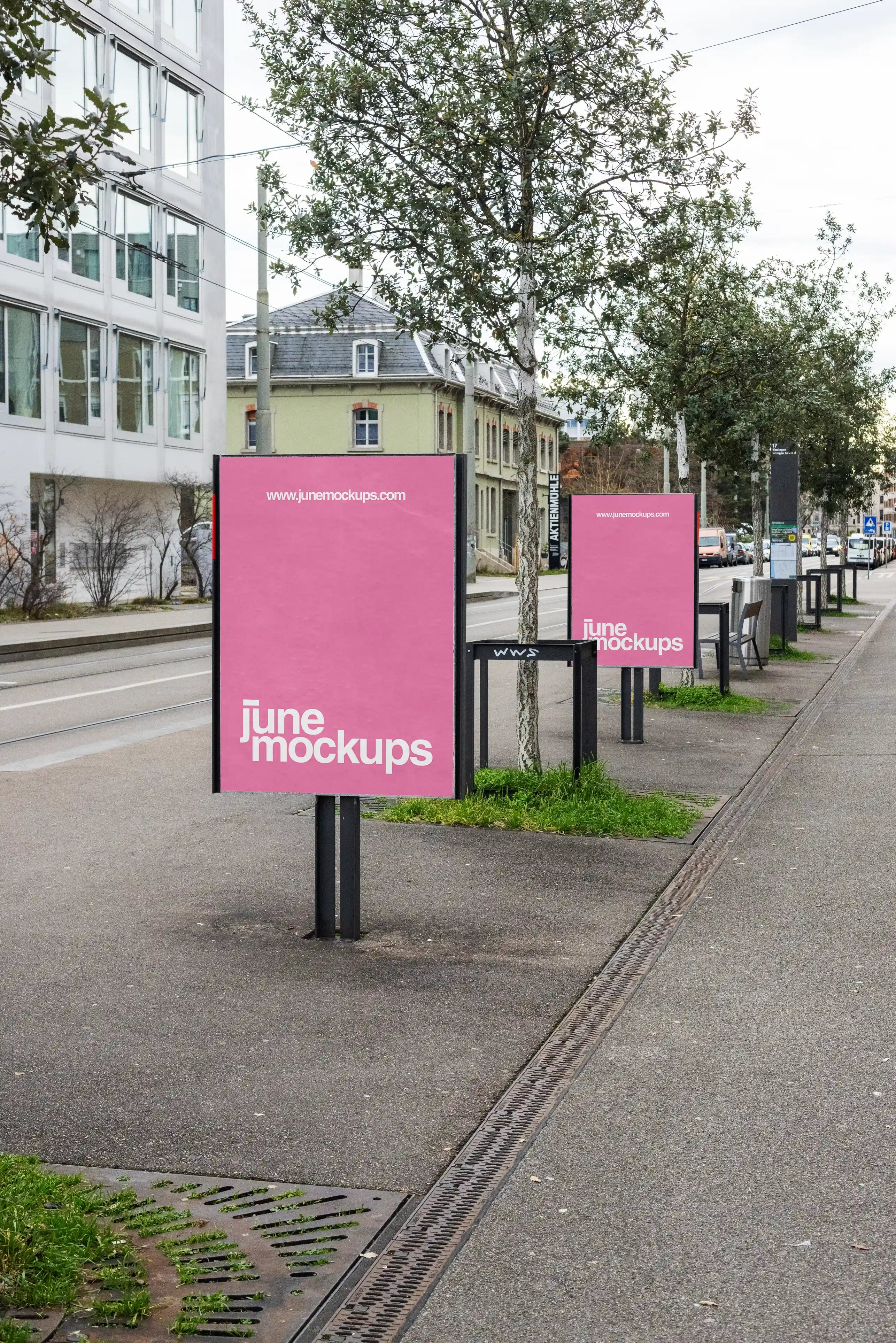Two pink DIN poster mockups displayed on freestanding outdoor poster boards at Wiesenplatz in Basel. The signs stand along a sidewalk with trees, tram tracks, grass patches, street furniture and surrounding urban buildings in the background.