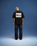Black oversized t-shirt mockup back view featuring large rectangular back print design, realistic fabric folds and shadows, worn by male model in clean studio environment with soft blue background, high resolution apparel mockup for streetwear branding and graphic design presentation.