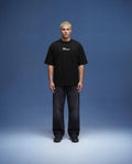Full body black oversized t-shirt mockup front view worn by male model with relaxed fit, realistic fabric drape and natural shadows, clean studio setup with blue background, high resolution apparel mockup for fashion brands, streetwear collections and e-commerce presentation.