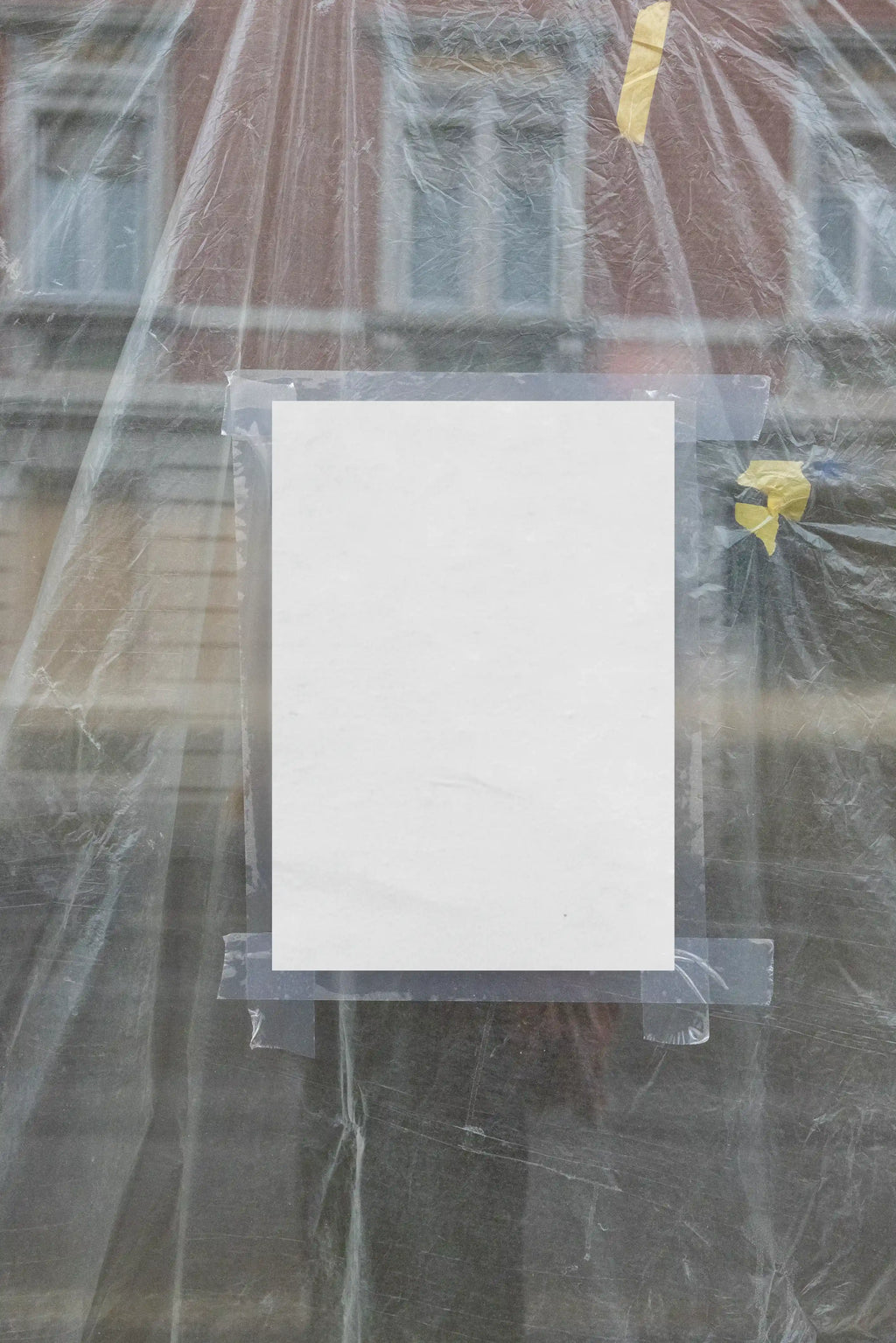 Blank white A4 poster mockup placed behind wrinkled transparent plastic foil on a window, held with visible tape and surrounded by reflections, soft grey tones and raw material textures, perfect for realistic flyer, poster and urban print design previews.