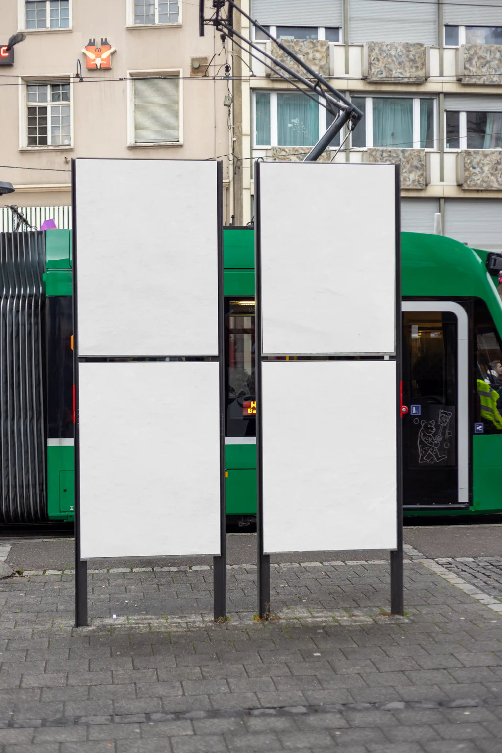 Four blank DIN poster mockups on two freestanding street advertising displays at Claraplatz in Basel, with a green tram, city architecture and urban pavement in the background. Ideal for clean poster series, campaign layouts and public transport advertising previews.